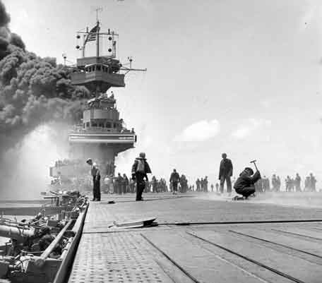 Pemandangan di kapal USS Yorktown (CV-5), tak lama setelah dia dihantam oleh tiga bom Jepang pada 4 Juni 1942. Asap tebal berasal dari kebakaran di serapannya, yang disebabkan oleh bom yang menusuk mereka dan merobohkan ketelnya. Pria dengan palu di kanan mungkin menutupi lubang masuk bom di lift depan. Perhatikan kabel gigi penahan dan elemen palisade depan di dek penerbangan; Antena radar CXAM, panji nasional besar, dan antena suar pelacak YE di atas tiang depan; Meriam kaliber 5 inci/38, kaliber .50, dan 1,1 inci berawak dan siap di kiri. Foto diambil oleh Fotografer Kelas 2 William G. Roy dari sisi kanan dek penerbangan, tepat di depan galeri senjata 5/38 depan (80-G-312018).